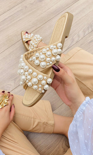 GRETA MODELO AGARRA SANDALIAS DE PISO CON PERLAS EN LA MANO CON LOOK DE PANTALON BEIGE Y BLUSA BLANCA PARA LLEVAR A LA PLAYA