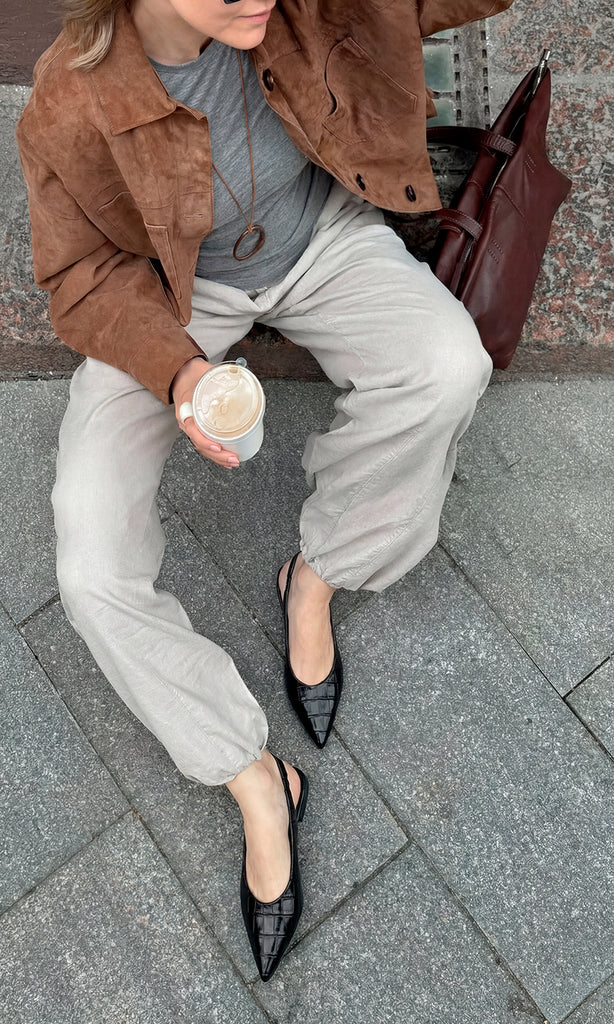 MELISSA CHICA COMBINA FLATS NEGROS ACABADO CROC CON PANTALON COLOR ARENA Y CAZADORA CAFE ENCIMA DE T-SHIRT GRIS MIENTRAS SOSTIENE UN LATTE