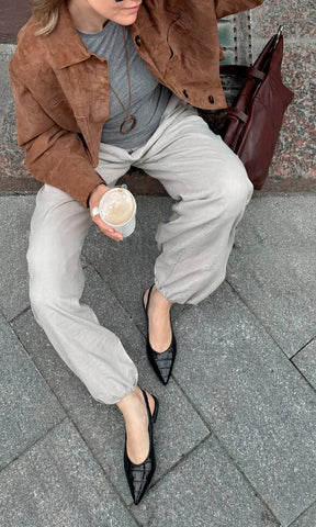 MELISSA CHICA COMBINA FLATS NEGROS ACABADO CROC CON PANTALON COLOR ARENA Y CAZADORA CAFE ENCIMA DE T-SHIRT GRIS MIENTRAS SOSTIENE UN LATTE