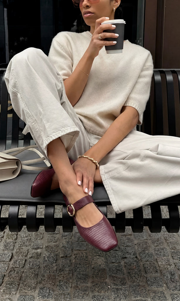 VALERIE MODELO POSA EN BANCA NEGRA FLATS COLOR BURGUNDY CON PANTALON BLANCO Y SUETER MANGA CORTA MIENTRAS SOSTIENE VASO DE CAFE