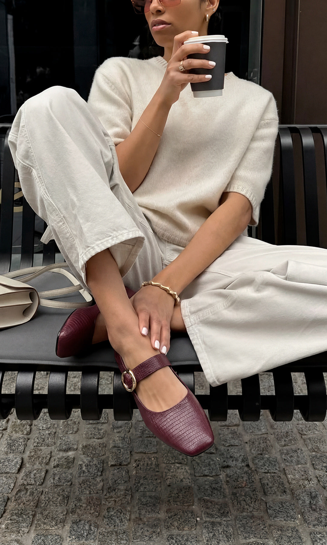 VALERIE MODELO POSA EN BANCA NEGRA FLATS COLOR BURGUNDY CON PANTALON BLANCO Y SUETER MANGA CORTA MIENTRAS SOSTIENE VASO DE CAFE