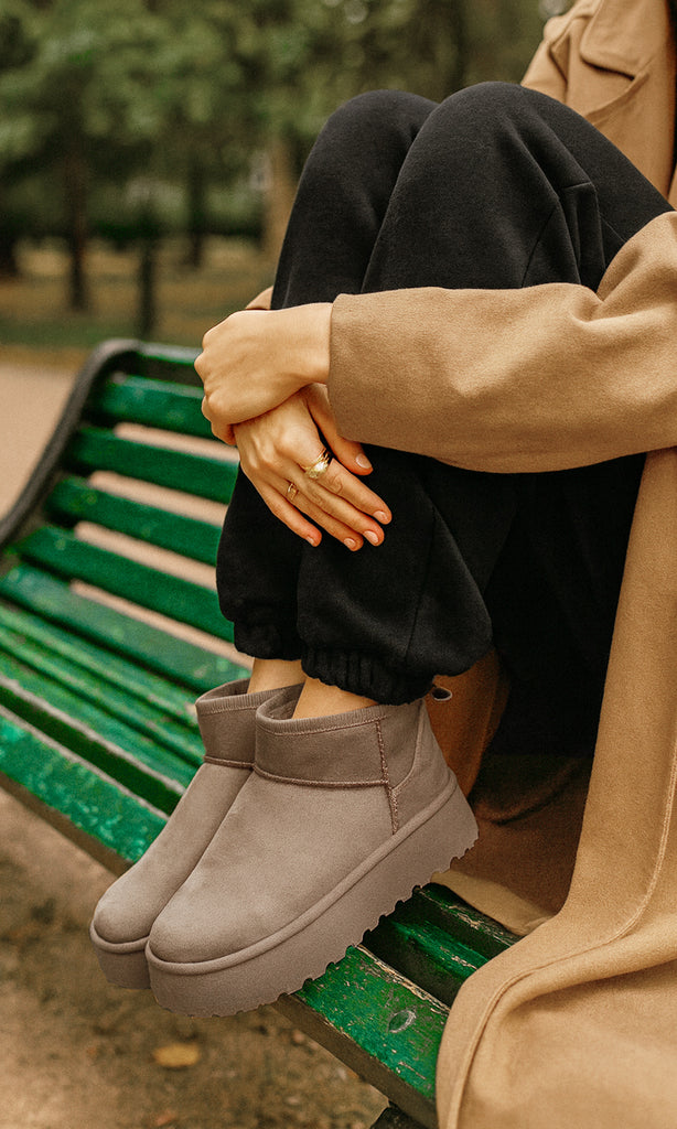 WINTER MUJER SENTADA EN BANCA VERDE COMBINANDO BOTAS CAMEL DE SUELA GRUESA CON PANTS NEGROS Y ABRIGO CAMEL PARA DÍA  AL AIRE LIBRE