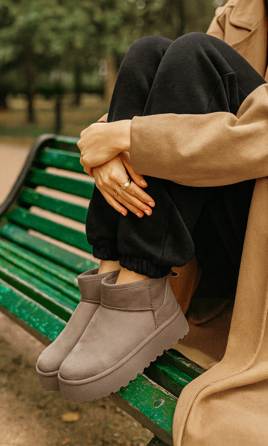WINTER MUJER SENTADA EN BANCA VERDE COMBINANDO BOTAS CAMEL DE SUELA GRUESA CON PANTS NEGROS Y ABRIGO CAMEL PARA DÍA  AL AIRE LIBRE