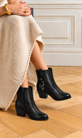Botines negros para mujer con tacón bloque, punta estilizada y detalle lateral colgante con aplicaciones doradas, usados con vestido tejido color marfil