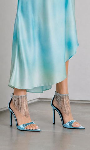 Sandalias ALESSANDRA color azul metálico con tacón de aguja, punta cuadrada y flecos de cristales en el tobillo, combinadas con vestido satinado para un look de invitada a boda.