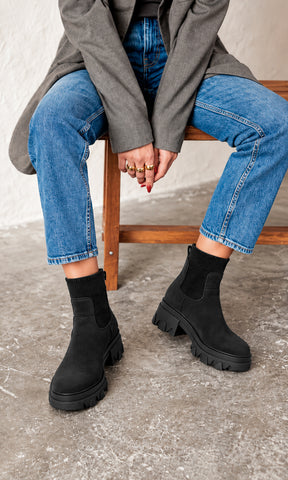 Botines Ashton negros estilo Chelsea con caña elástica de punto, suela track robusta y tacón chunky bajo, combinados con jeans azules y blazer gris en un look casual urbano