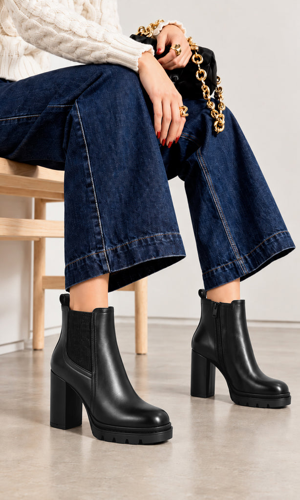 Botines negros para mujer estilo Chelsea con panel elástico lateral, cierre interno, tacón alto de bloque y suela track, combinados con jeans de pierna ancha y suéter de punto blanco en un look casual contemporáneo
