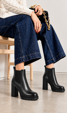 Botines negros para mujer estilo Chelsea con panel elástico lateral, cierre interno, tacón alto de bloque y suela track, combinados con jeans de pierna ancha y suéter de punto blanco en un look casual contemporáneo