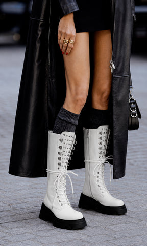 Botas Epic blancas estilo combat con plataforma chunky, agujetas y estoperoles metálicos, combinadas con abrigo negro largo en un look urbano de alto contraste