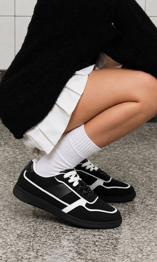 Sneakers Matcha negros de inspiración retro, con perfil bajo, paneles y detalles blancos en contraste, cordones blancos y suela plana oscura, usados con calcetines y una falda corta