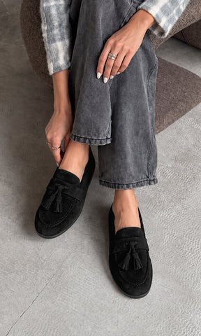 Mocasines negros con borlas y acabado tipo gamuza, combinados con jeans grises de pierna ancha y suéter tejido para un look casual para mujer