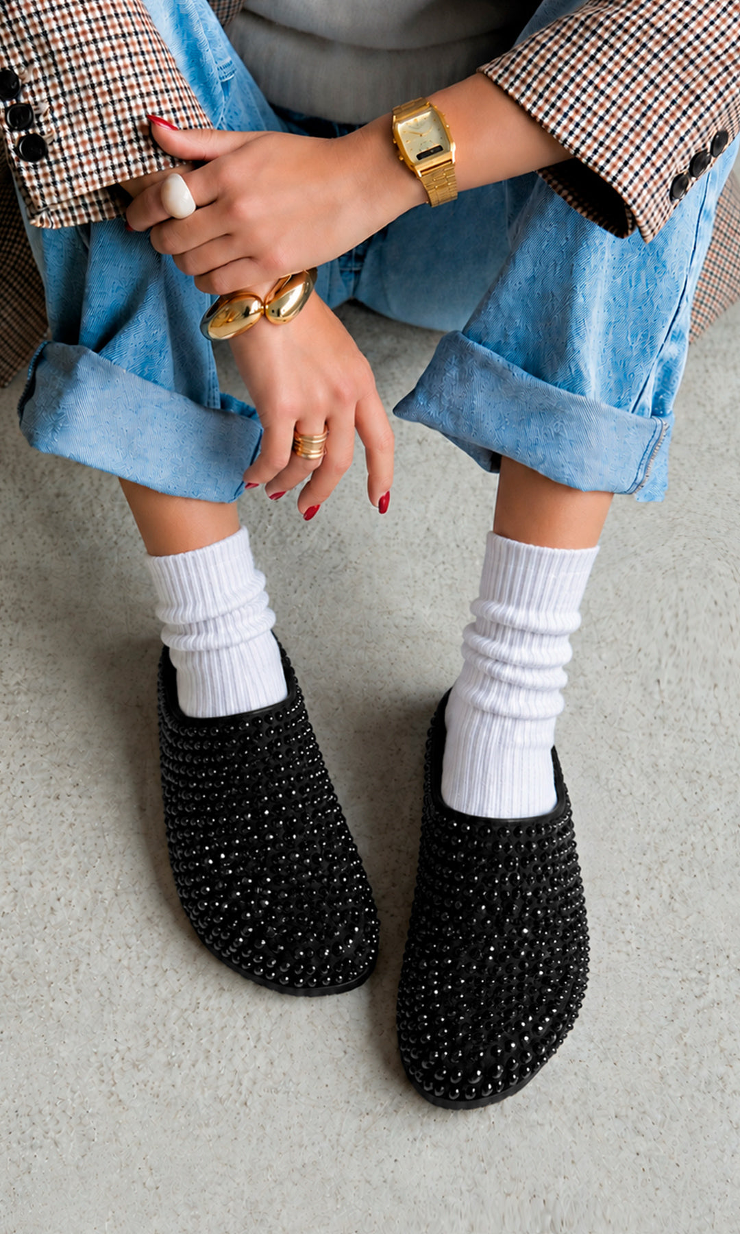 Mules negros planos cubiertos de pedrería brillante, combinados con calcetas blancas, jeans claros y blazer de cuadros en un look casual moderno