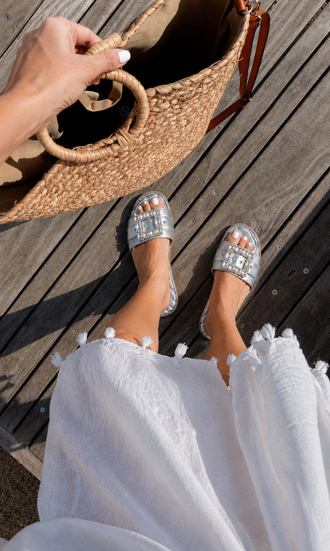 Sandalias planas PRISCILA plateadas con maxi hebilla de pedrería, combinadas con vestido blanco y bolso de rafia para un look resort sofisticado