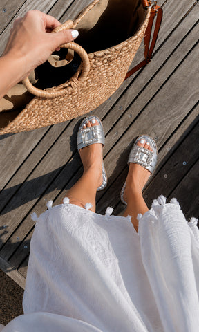 Sandalias planas PRISCILA plateadas con maxi hebilla de pedrería, combinadas con vestido blanco y bolso de rafia para un look resort sofisticado