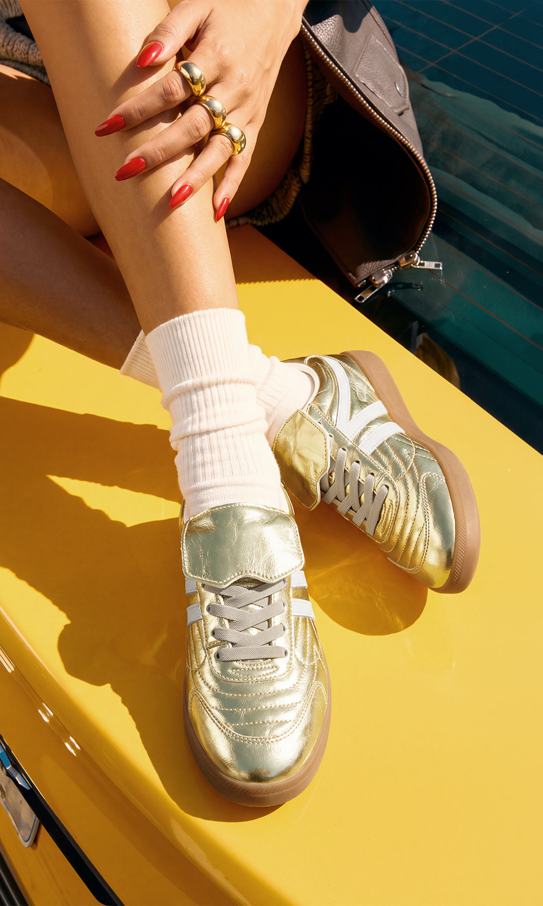 Tenis dorados metálicos para mujer con agujetas blancas, franjas laterales blancas y suela color caramelo, combinados con calcetas blancas sobre un automóvil amarillo