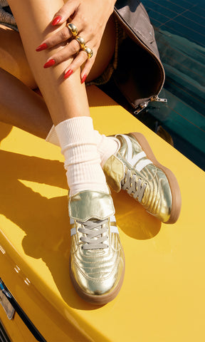 Tenis dorados metálicos para mujer con agujetas blancas, franjas laterales blancas y suela color caramelo, combinados con calcetas blancas sobre un automóvil amarillo