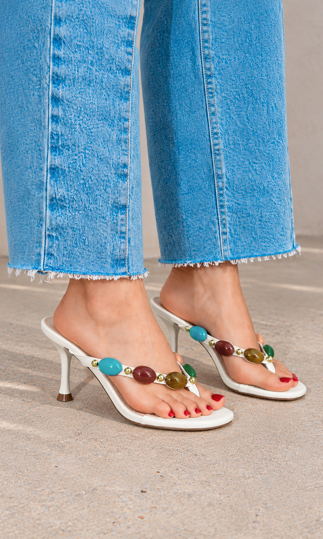 Sandalia Tribeca blanca estilo mule, con tacón fino medio, punta abierta y tira delantera decorada con piedras multicolor y detalles dorados, combinada con jeans azules de bajo desflecado
