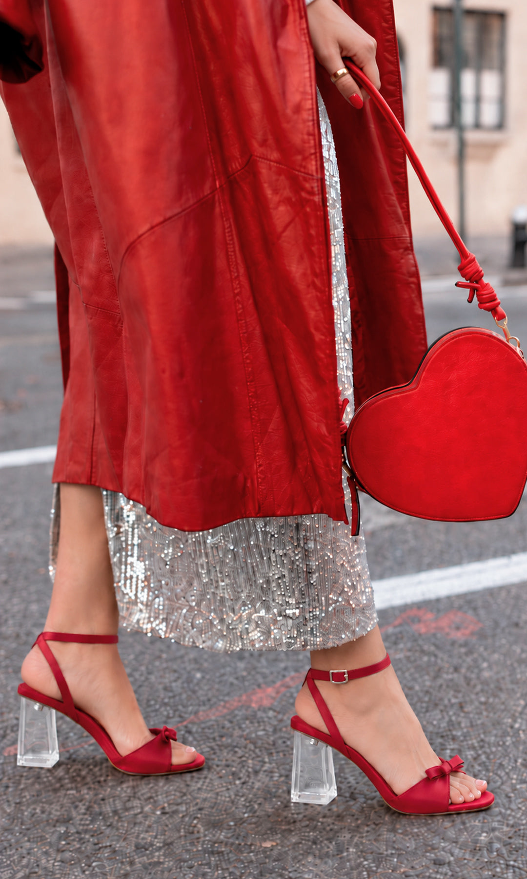 Zapatillas rojas con tacón ancho transparente, pulsera al tobillo y detalle de moño, combinadas con abrigo rojo, falda plateada de lentejuelas y bolso de corazón en un look de fiesta