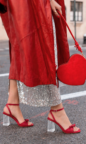 Zapatillas rojas con tacón ancho transparente, pulsera al tobillo y detalle de moño, combinadas con abrigo rojo, falda plateada de lentejuelas y bolso de corazón en un look de fiesta